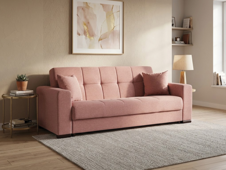 Pink upholstered sofa with two matching cushions in a cozy living room setting, beige walls, and a side table with a plant to the left.