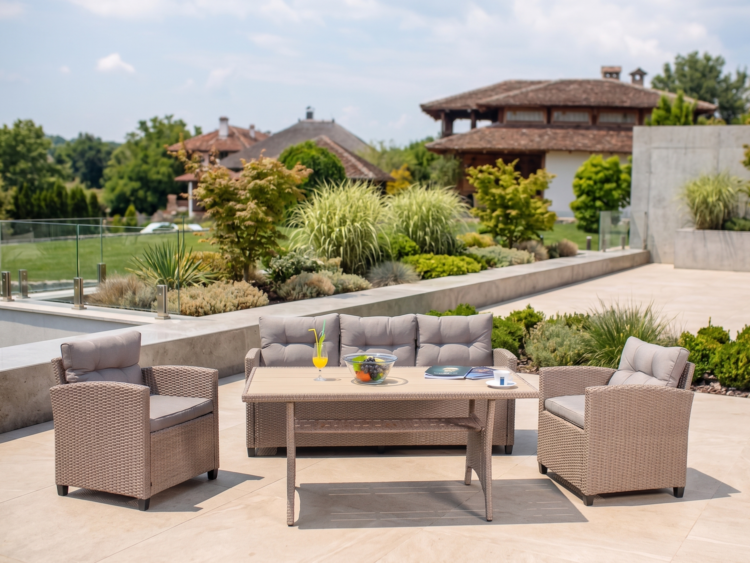 Wicker outdoor seating area on a sunny patio: sofa and two armchairs around a rectangular table with a fruit bowl, a yellow drink, magazines, and a cup, with landscaped garden and houses in the background.