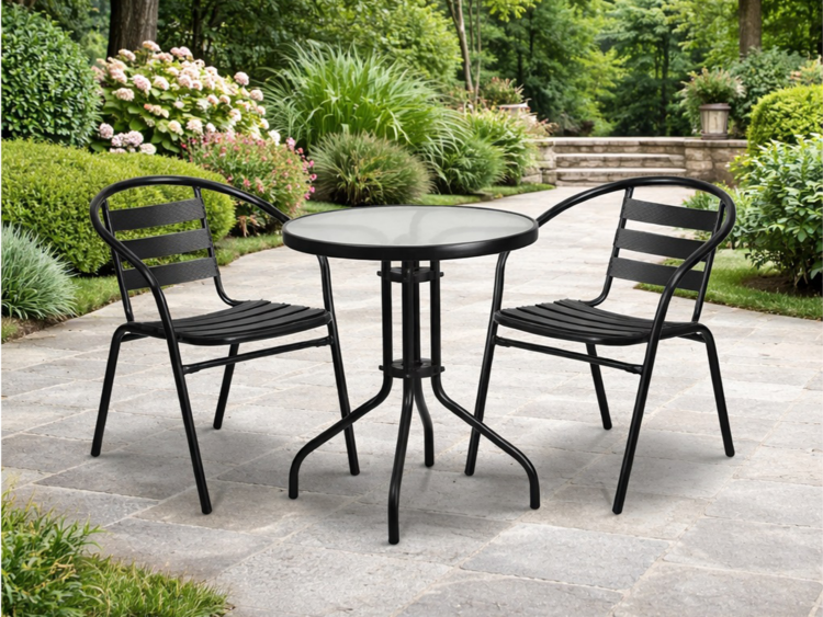 Outdoor patio scene with a round glass-top table and two black metal chairs on a stone-tiled patio, surrounded by lush greenery and flowering shrubs.