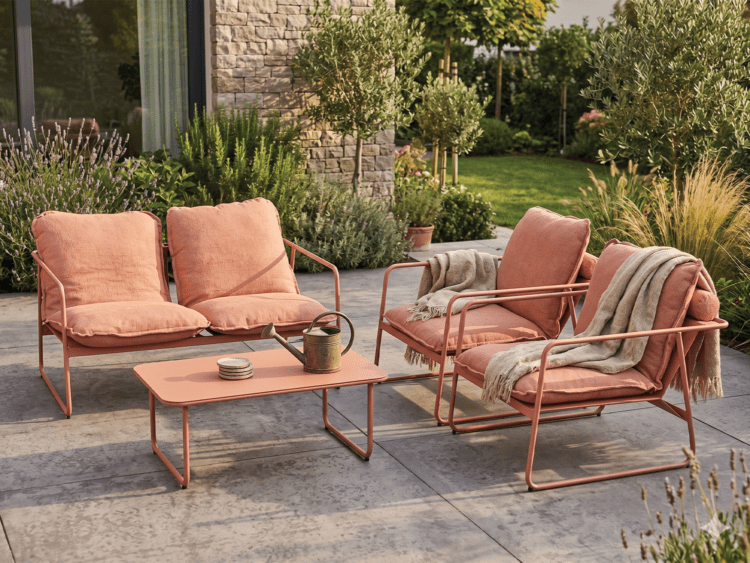 Peach-colored metal outdoor seating set with a two-seat sofa, two armchairs, a matching coffee table, and blankets on the chairs in a landscaped courtyard patio.