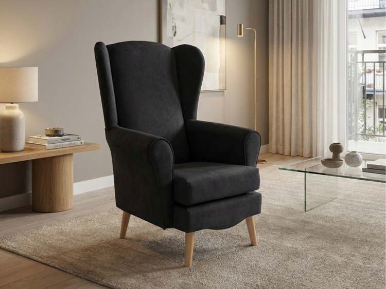 Dark upholstered wing chair with light wood legs sits on a beige textured rug beside a wooden side table with a lamp in a modern living room.