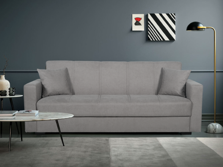 Gray upholstered sofa with two matching cushions in a modern living room, flanked by a round marble coffee table and a black-and-gold floor lamp against a blue-gray wall with abstract art.