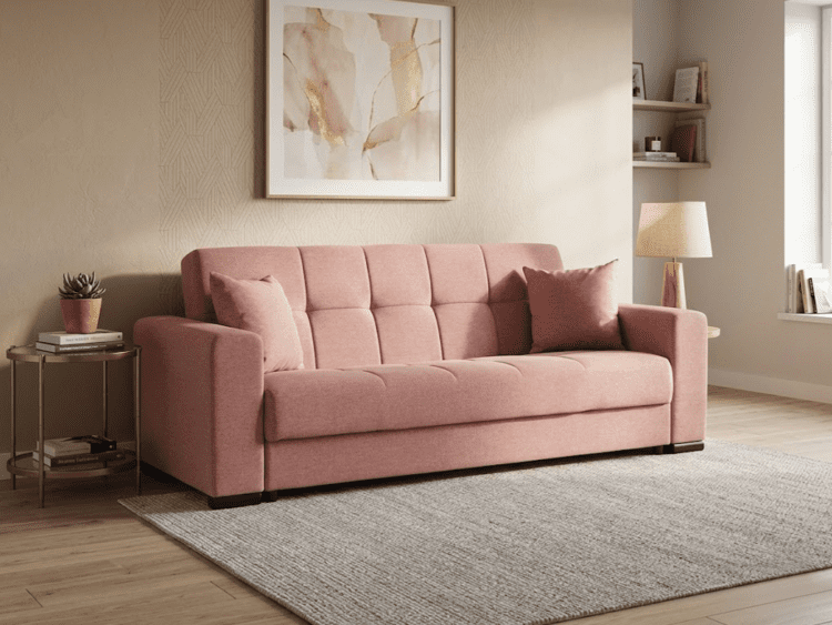 Pink upholstered sofa with two matching cushions in a cozy living room setting, beige walls, and a side table with a plant to the left.