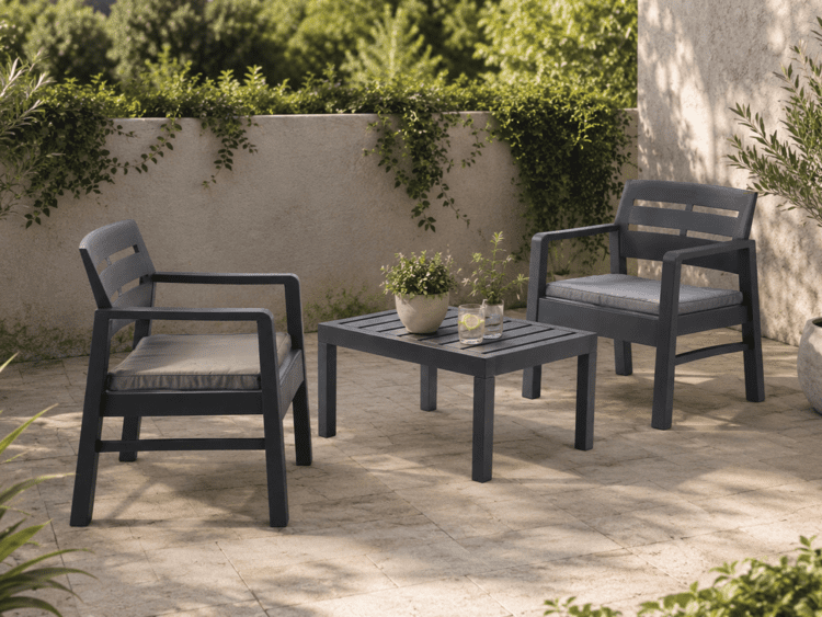 Two dark gray outdoor armchairs with cushions around a matching square table on a stone patio, a potted plant and two drinks on the table